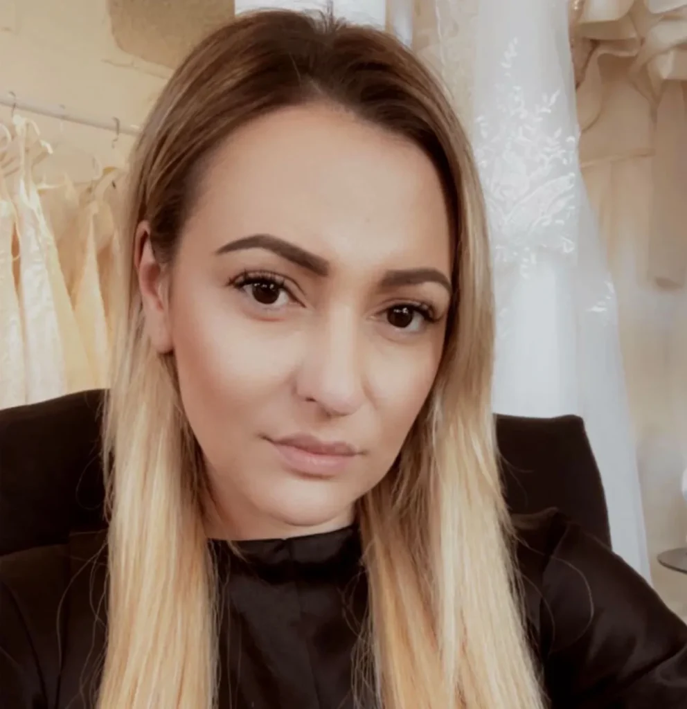Woman with long blonde hair in a black shirt, in front of fabric or clothes on hangers, looking at the camera.