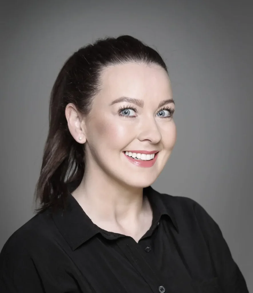 Smiling woman with dark hair in a ponytail wearing a black shirt, against a gray background.