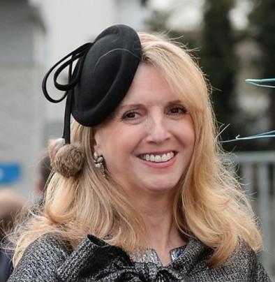 A woman smiling, wearing a black hat with a pom-pom, in an outdoor setting.