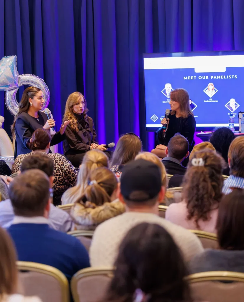 Panel discussion at a conference with speakers engaging a seated audience, event screen in the background.