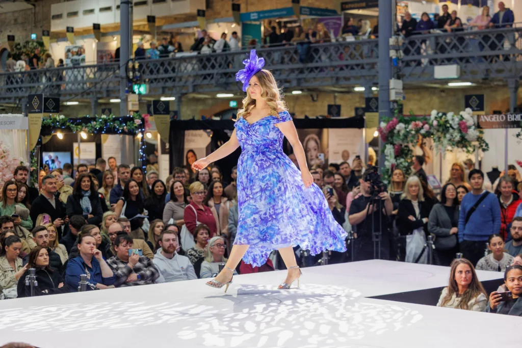 Model in a blue floral dress and hat walking a runway at a fashion show with audience watching.