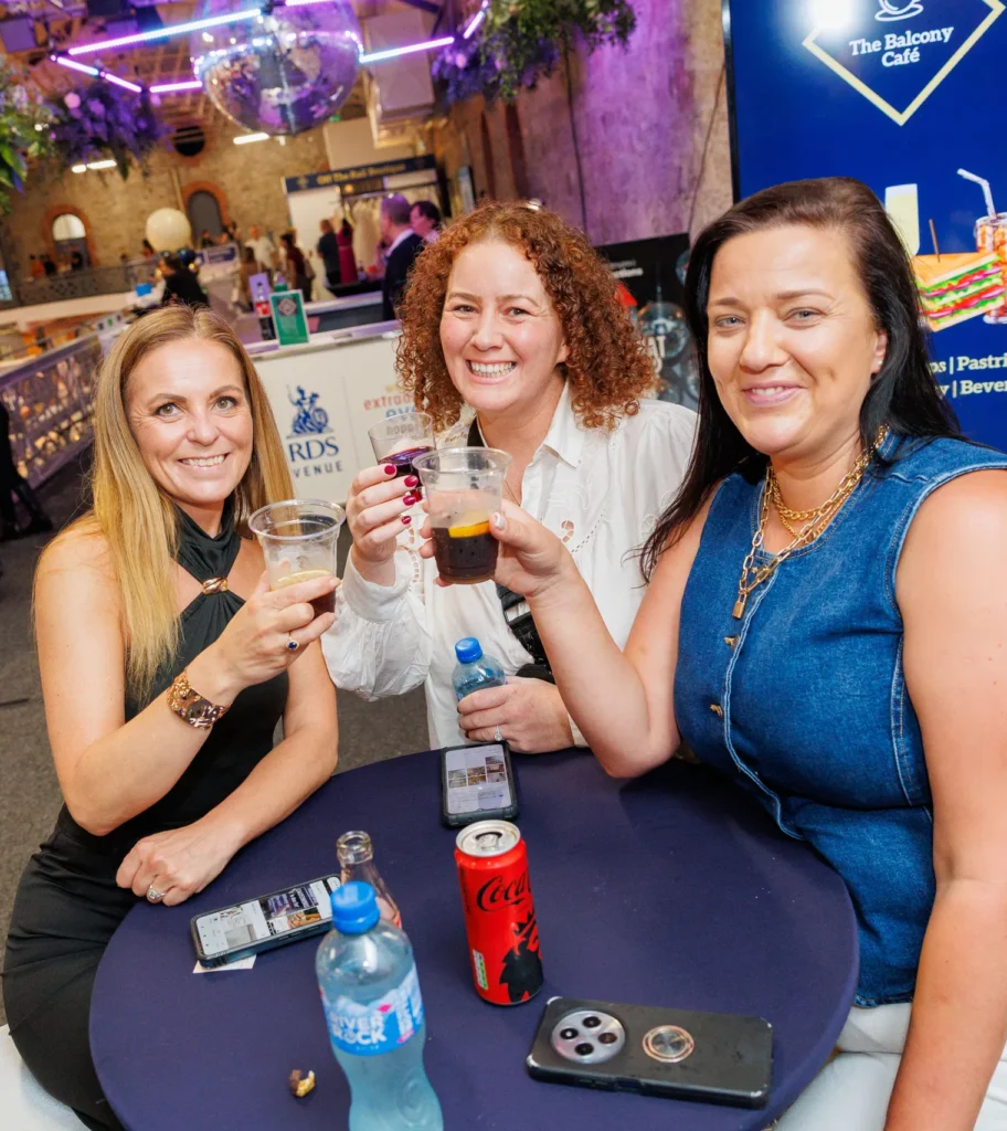 Three women enjoying drinks and socializing at a lively indoor event with colorful decor and a cafe sign in the background.