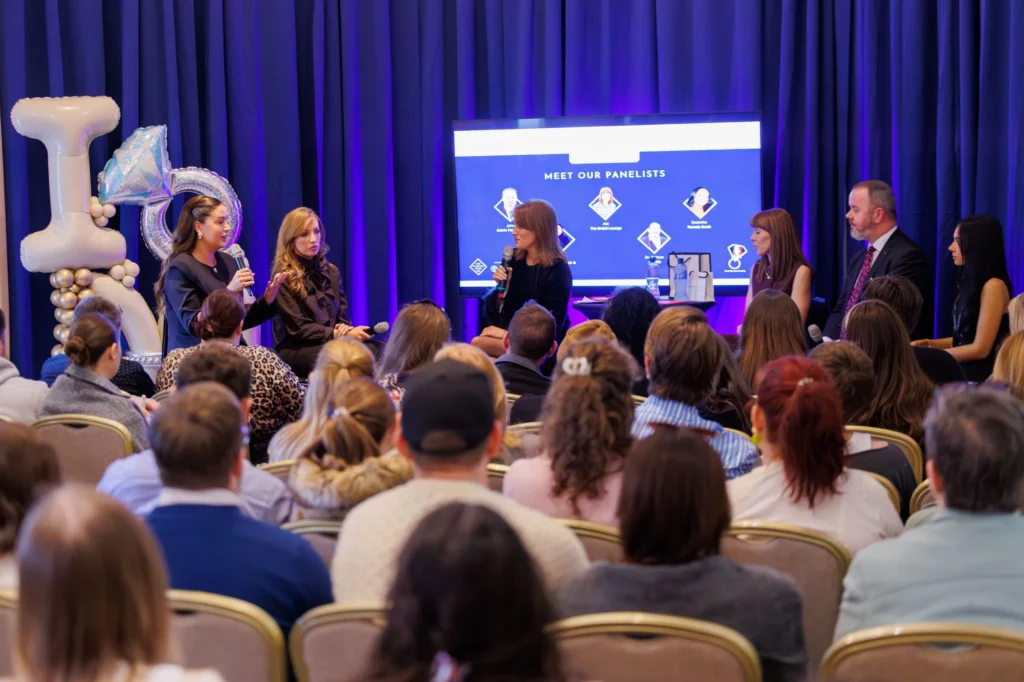 Panel discussion at a conference with speakers and audience, blue background, balloons, and screen display.