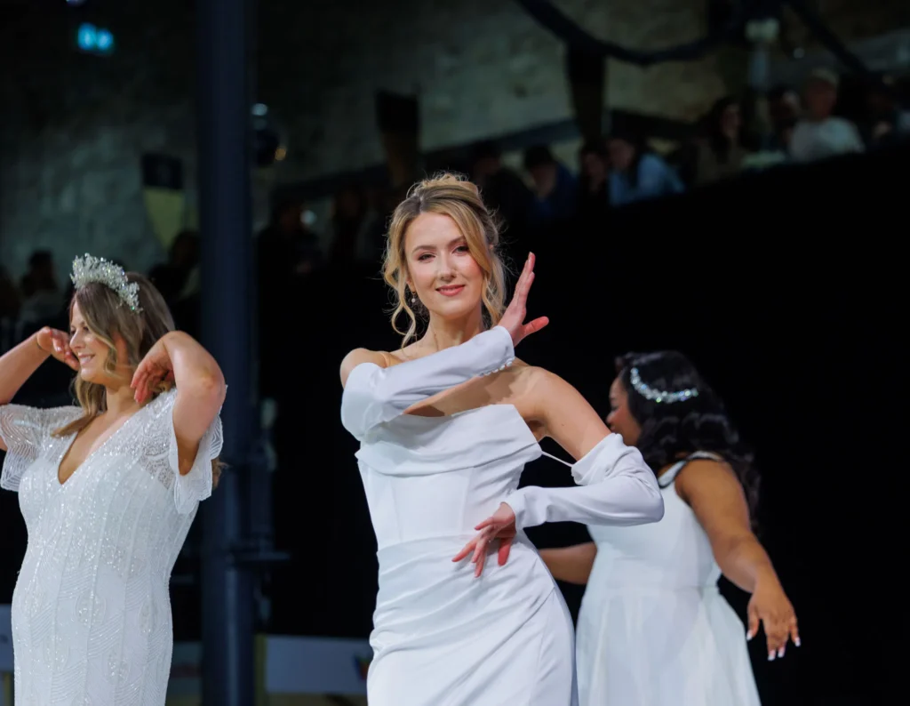 Models in white gowns dance gracefully on a fashion show runway.