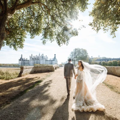 Bride and groom walking near a castle under sunny skies, surrounded by nature.