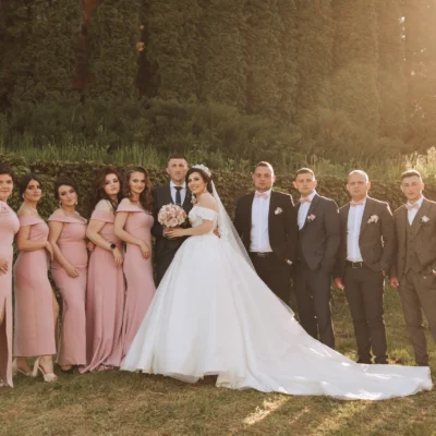 Wedding party poses in elegant attire, with bridesmaids in pink and groomsmen in suits, amid lush greenery.