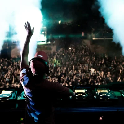 DJ performing live at a concert with vibrant lights and an energetic crowd in the background.